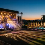 An outdoor festival stage with production lighting and a poolside hotel lounge area bathed in warm and cool tones at dusk