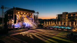 An outdoor festival stage with production lighting and a poolside hotel lounge area bathed in warm and cool tones at dusk