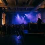 Industrial concrete venue interior lit by deep blue and purple stage lights, with distant blurred crowd and professional DJ equipment