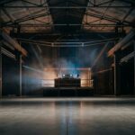 Empty warehouse hall with steel trusses and a raised DJ booth holding a mixer and media players, lit by blue and amber beams through haze.