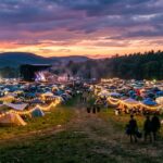 Festival campground at sunset with illuminated tents, distant stage, string lights, and atmospheric haze creating an immersive electronic music event atmosphere.