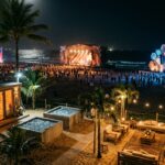 Festival stages and wellness area on a Brazilian beach at night, with distant crowd silhouettes and ocean backdrop