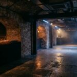Empty underground club with industrial vaulted ceiling, DJ booth with equipment, and atmospheric stage lighting reflecting off concrete floor